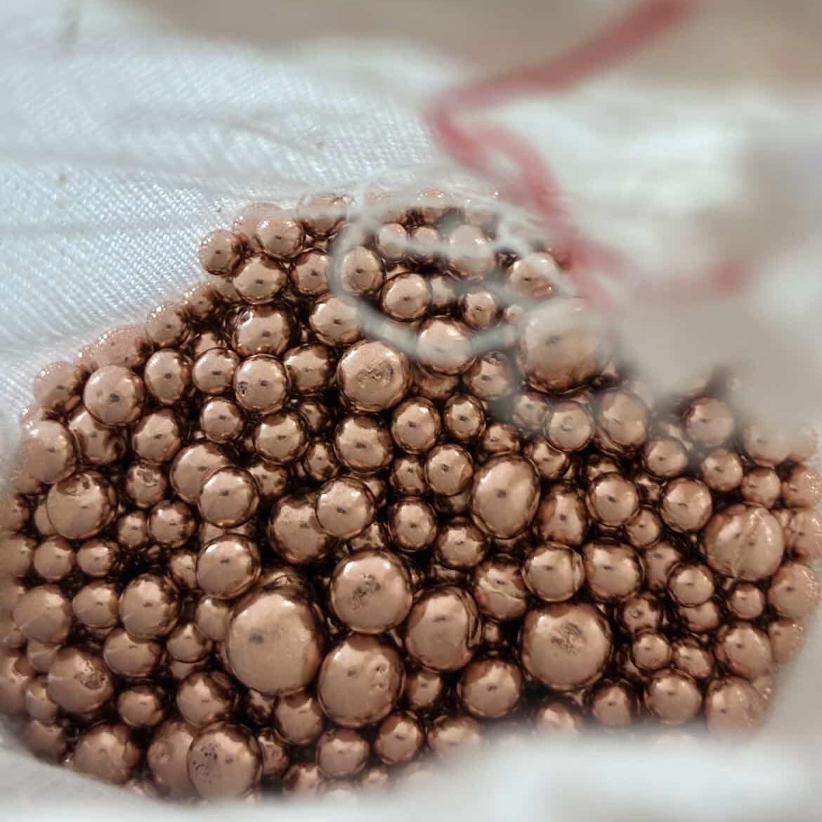 A close-up of a pile of small metallic copper-colored beads or granules resting on a white textured surface.