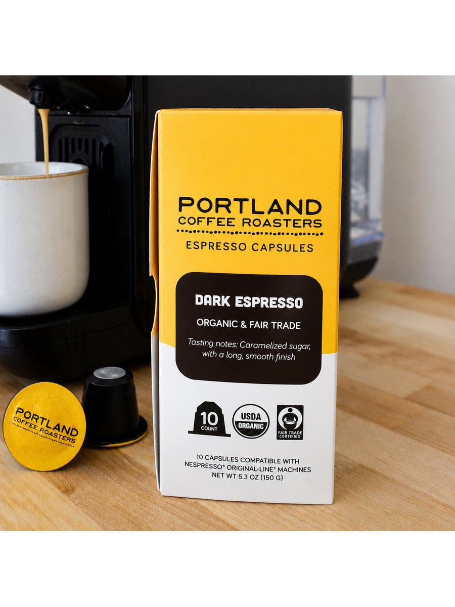 Portland Coffee Roasters Dark Espresso capsule box standing on a wooden countertop, with a coffee machine and a brewed cup in the background.