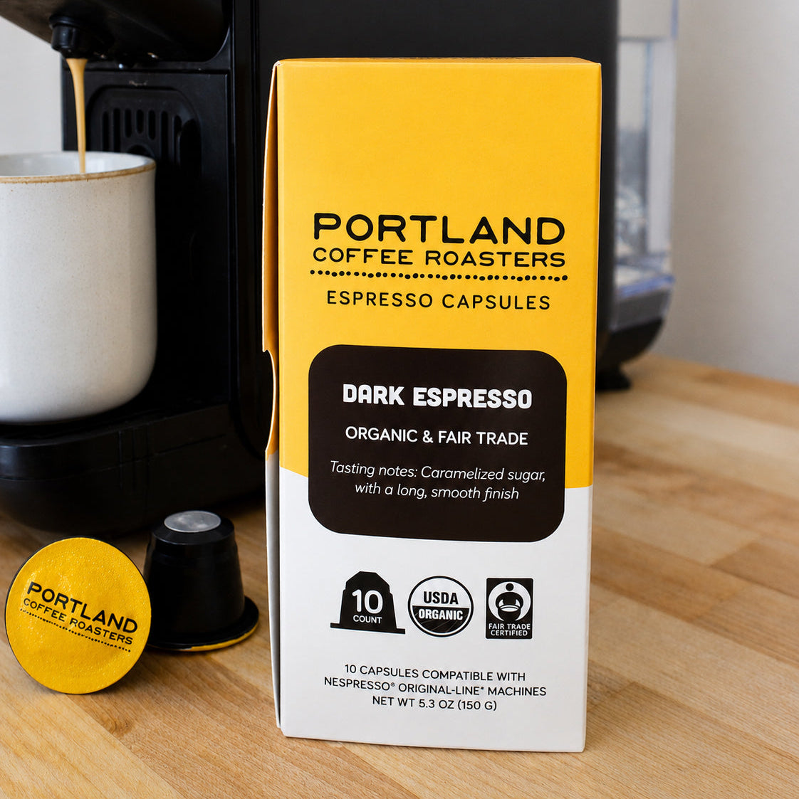 Portland Coffee Roasters Dark Espresso capsule box standing on a wooden countertop, with a coffee machine and a brewed cup in the background.