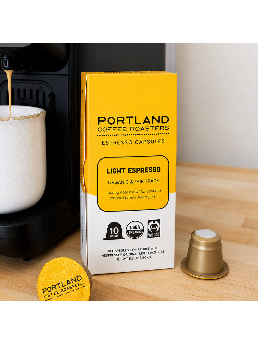 Portland Coffee Roasters Light Espresso capsule packaging box on a wooden countertop next to a coffee machine, with a loose gold capsule and a yellow capsule lid in front.