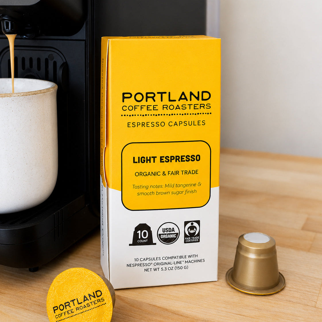 Portland Coffee Roasters Light Espresso capsule packaging box on a wooden countertop next to a coffee machine, with a loose gold capsule and a yellow capsule lid in front.