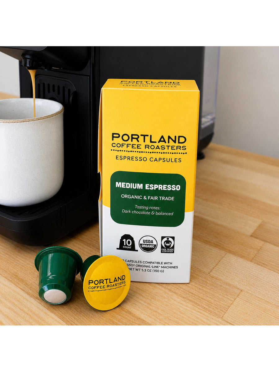 Portland Coffee Roasters Medium Espresso capsule box on a wooden table next to two capsules, one green and one yellow, with espresso flowing from a coffee machine into a white mug.