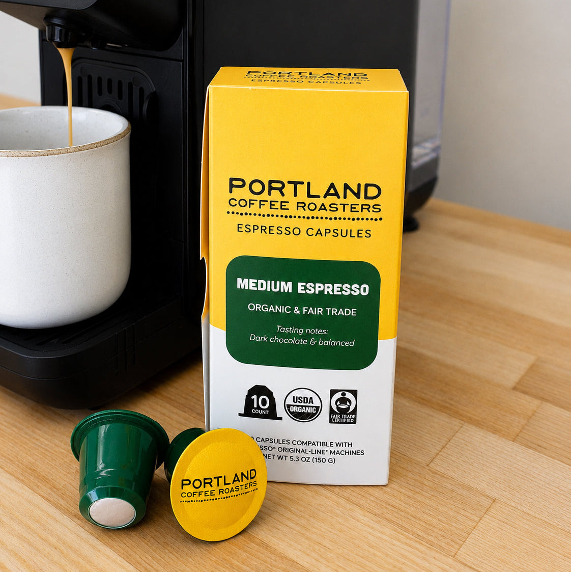 Portland Coffee Roasters Medium Espresso capsule box on a wooden table next to two capsules, one green and one yellow, with espresso flowing from a coffee machine into a white mug.