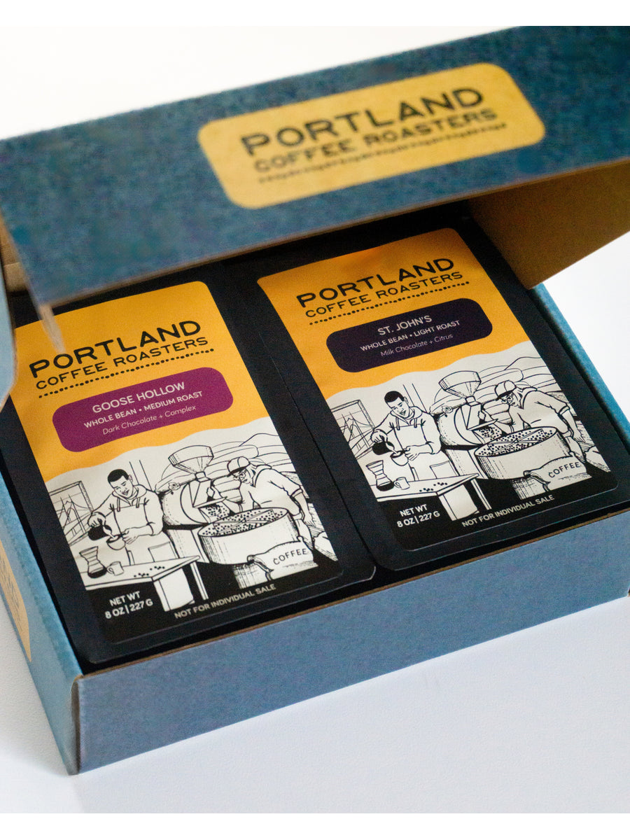 Opened blue cardboard box containing two black and yellow coffee bags from Portland Coffee Roasters. Each bag has an illustrated depiction of coffee preparation and flavor descriptions, with the bag on the left being Goose Hollow, Medium Roast, Dark Chocolate + Complex, and the bag on the right being St. John's, Light Roast, Milk Chocolate + Citrus.
