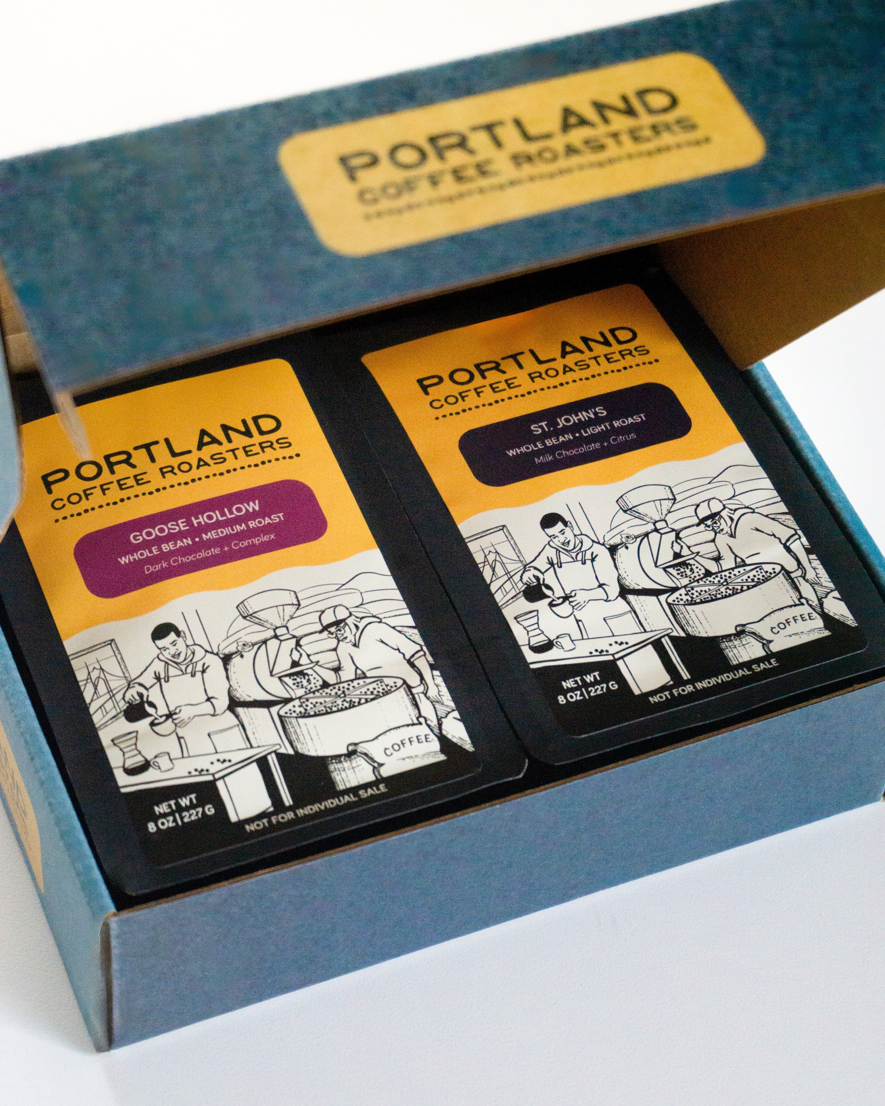 Opened blue cardboard box containing two black and yellow coffee bags from Portland Coffee Roasters. Each bag has an illustrated depiction of coffee preparation and flavor descriptions, with the bag on the left being Goose Hollow, Medium Roast, Dark Chocolate + Complex, and the bag on the right being St. John's, Light Roast, Milk Chocolate + Citrus.
