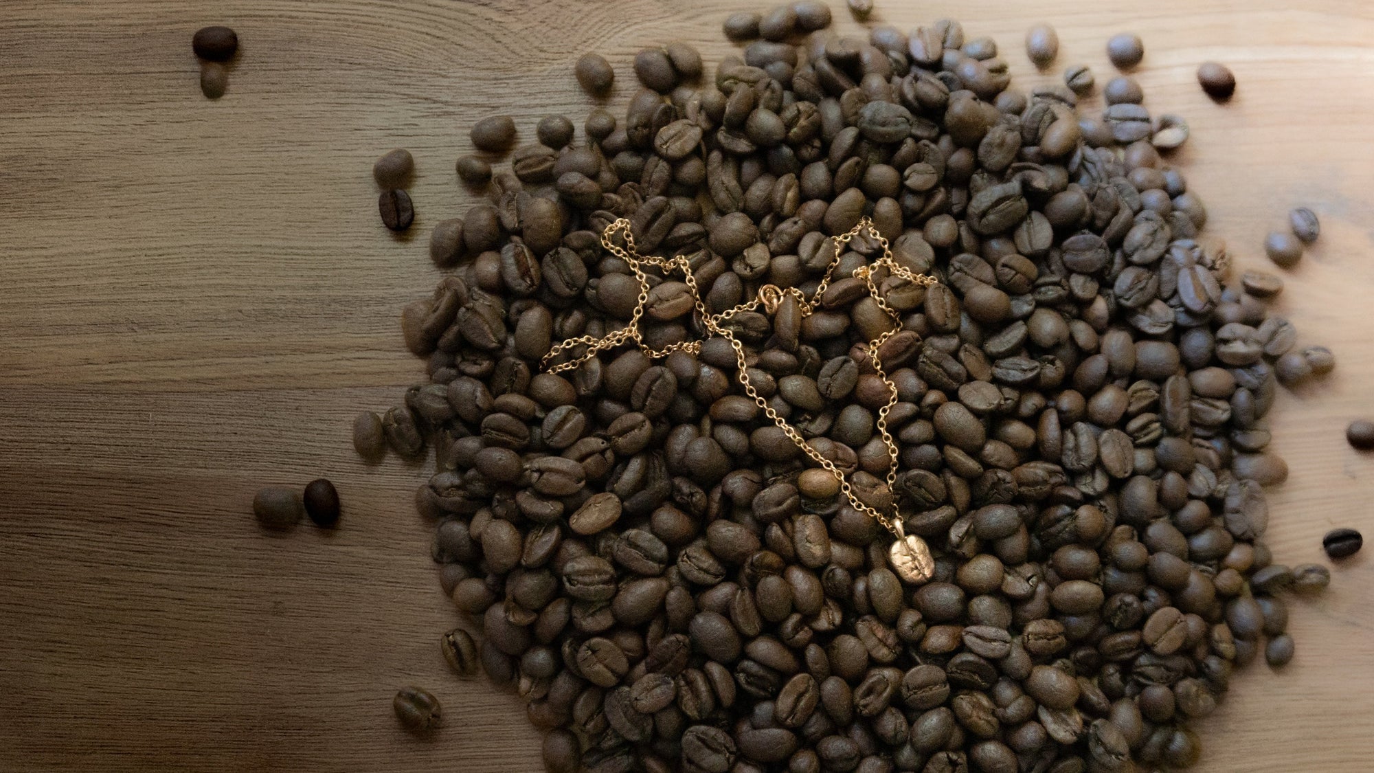 A gold chain necklace with a small pendant lies on top of a pile of roasted coffee beans on a wooden surface.