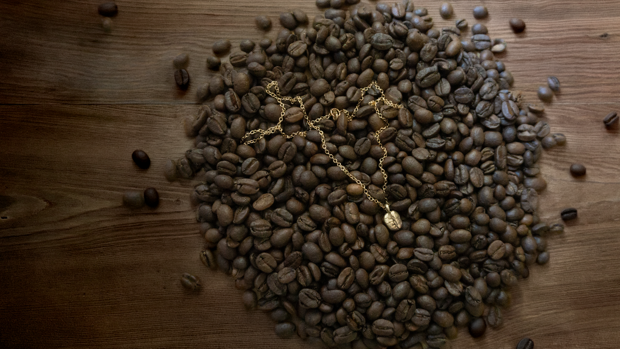 A gold-toned chain necklace with a small pendant lies on top of a pile of roasted coffee beans arranged on a rustic wooden surface.
