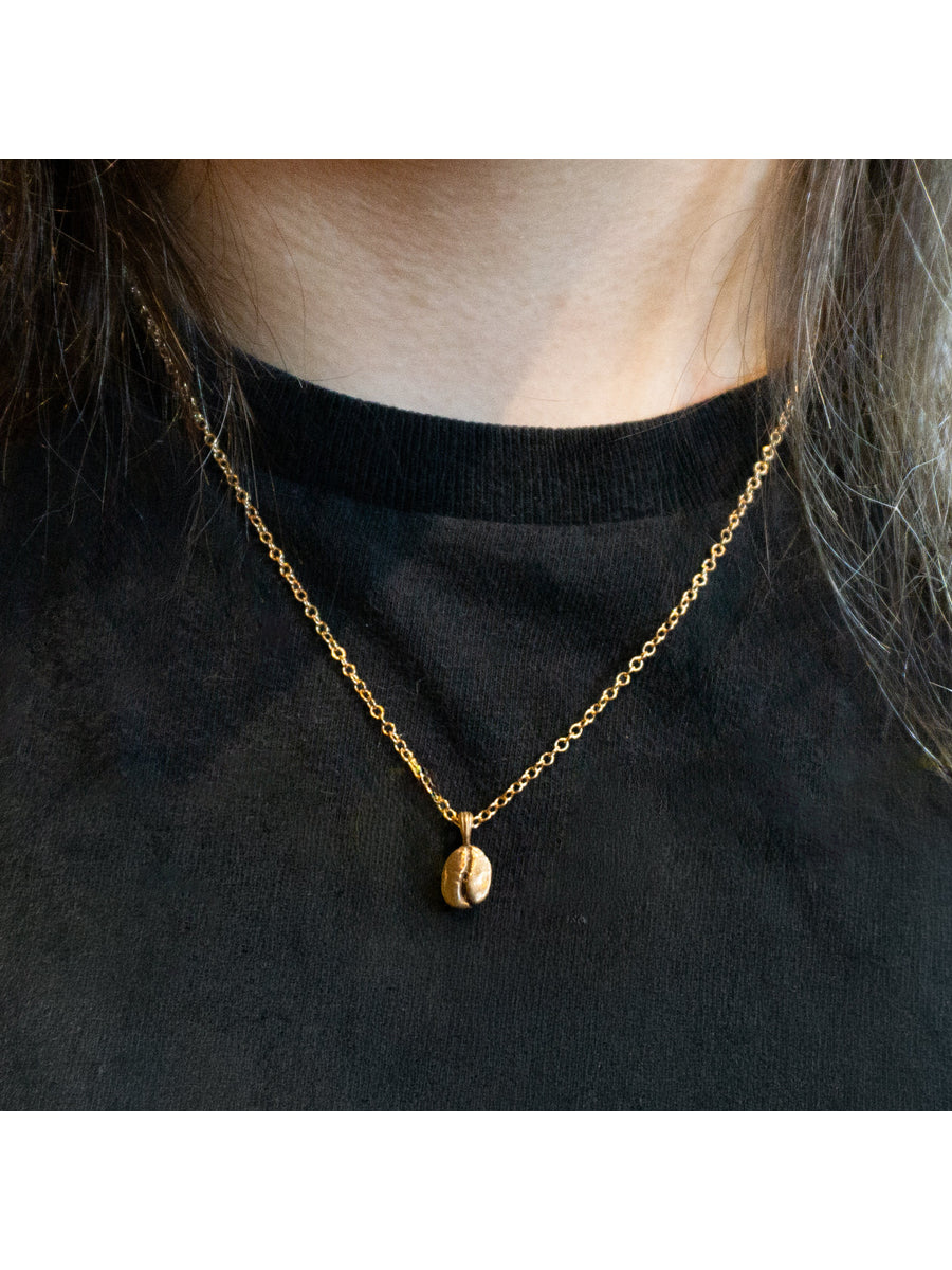 A close-up of a person wearing a delicate gold chain necklace with a small coffee bean pendant, styled over a black crewneck top.