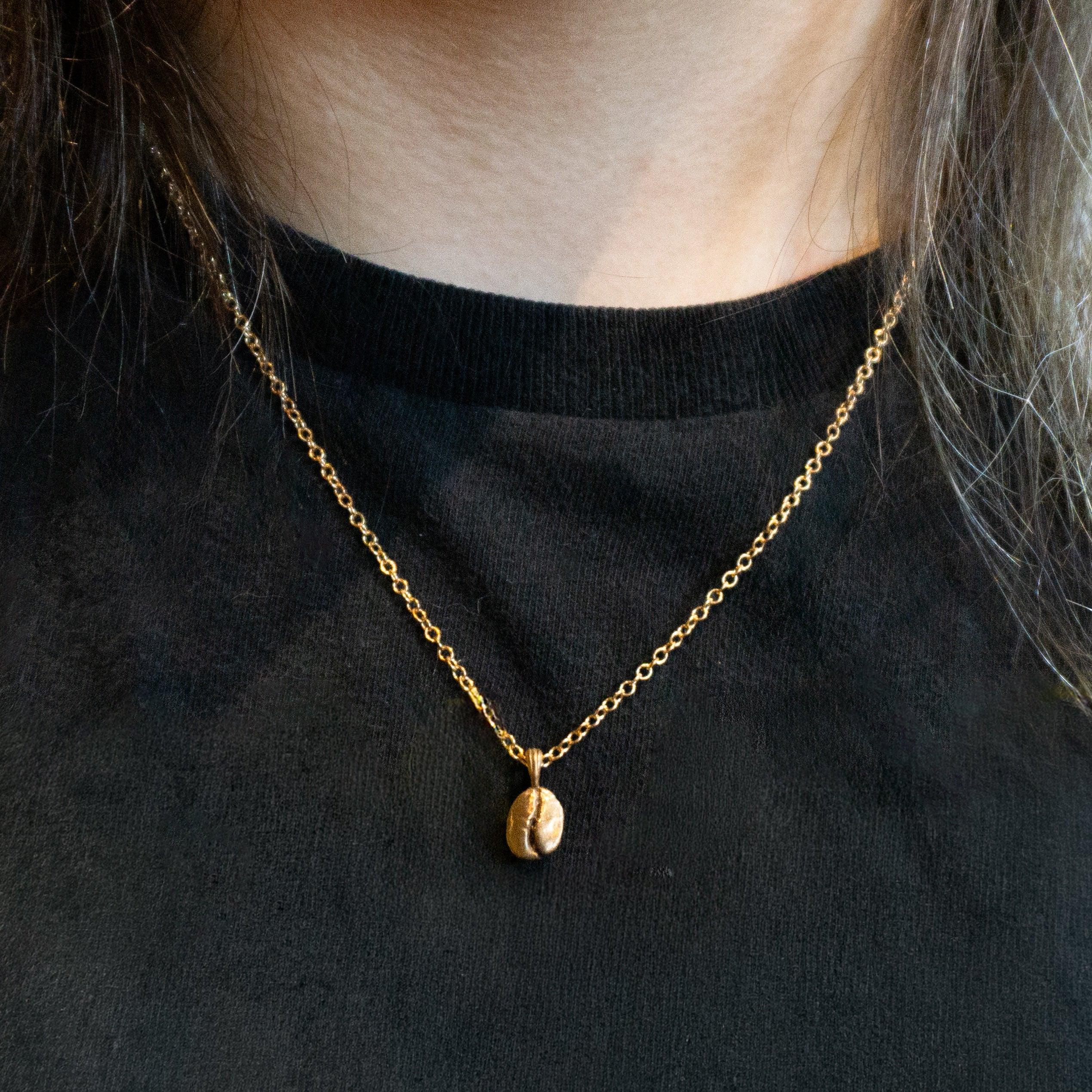 A close-up of a person wearing a delicate gold chain necklace with a small coffee bean pendant, styled over a black crewneck top.