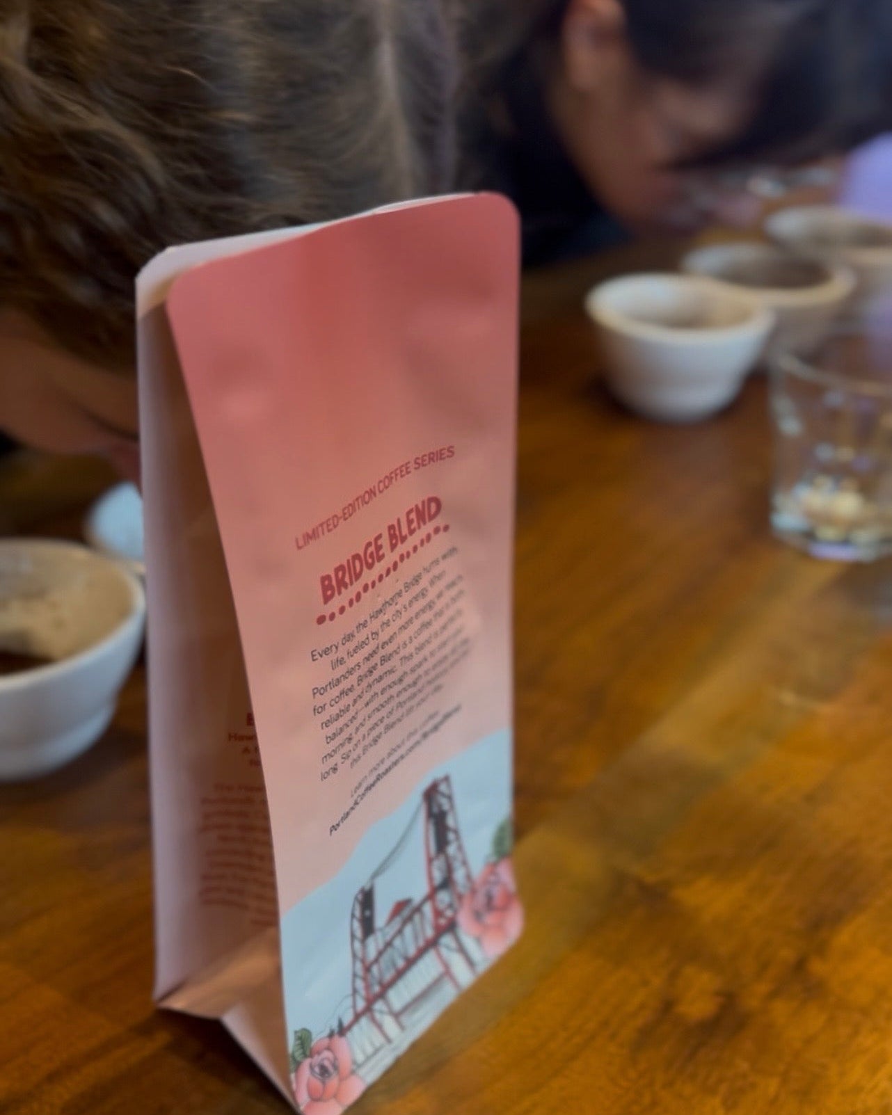 Two people sniffing coffee with a "Bridge Blend" bag in the foreground, small white bowls, and clear glasses on a wooden table.