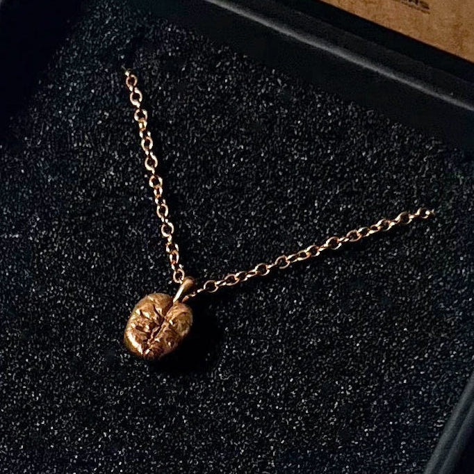 A gold necklace with a small textured heart pendant displayed inside an open black presentation box on a white fabric background.